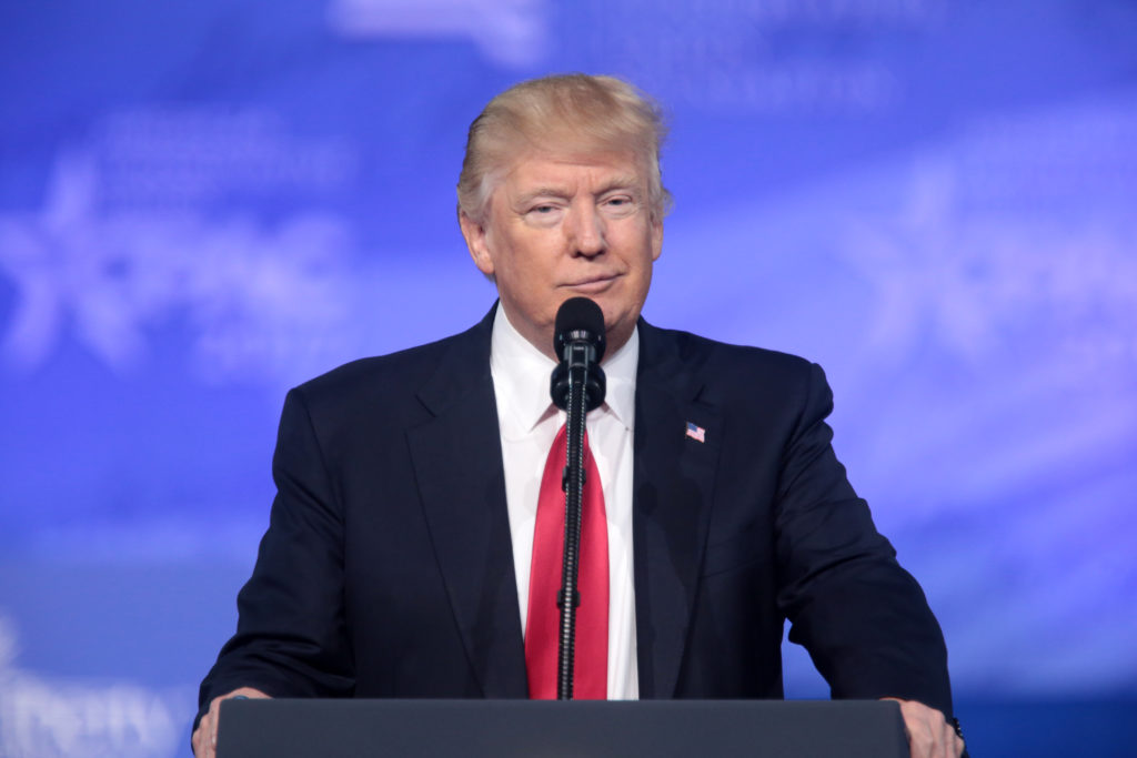 Uśmiechnięty Donald Trump stoi przy mównicy, w klapie ma wpięty znaczek z flagą Ameryki. W tle rozmyta ścianka z podpisem Conservative Political Action Conference (CPAC) in National Harbor. 