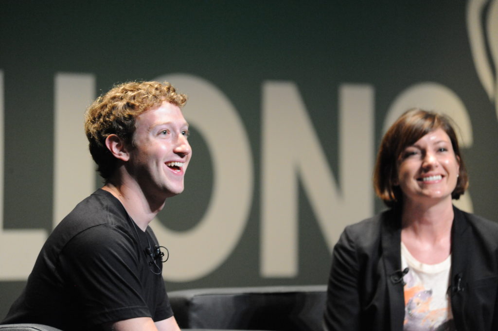 Uśmiechnięty Mark Zuckerberg podczas Cannes Lions 2010 siedzi na kanapie. Po prawej siedzi również uśmiechnięta kobieta.